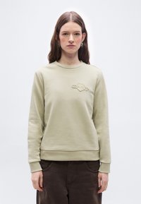 Νεαρή γυναίκα με μακριά καστανά μαλλιά, φορώντας ανοιχτό μπεζ φούτερ με λογότυπο στο στήθος και σκούρο καφέ παντελόνι, στέκεται μπροστά σε απλό φόντο.