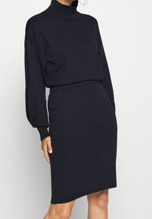 Donna che indossa un vestito nero aderente in maglia con maniche lunghe e collo alto, in piedi davanti a uno sfondo chiaro e uniforme.
