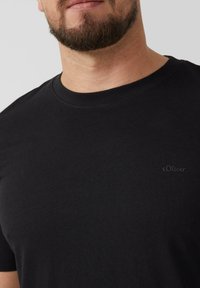 Zwarte katoenen T-shirt met ronde hals, voorzien van een subtiele s.Oliver-logo in donkere borduursel op de linkerkant van de borst. Glad textuur.
