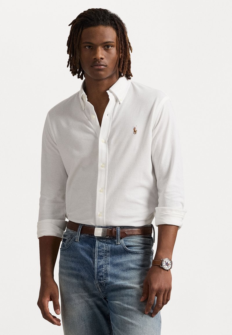White button-up shirt with long sleeves, paired with faded blue jeans and a brown leather belt. A wristwatch with a round face is visible.