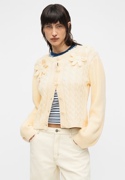 Γυναίκα που φοράει εκρού ζακέτα με πλεκτό σχέδιο και τρισδιάστατες λουλουδένιες λεπτομέρειες, ριγέ navy και λευκή κοντή μπλούζα, και ψηλόμεσο εκρού παντελόνι με τα χέρια στις τσέπες.