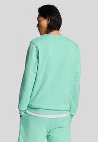 Sudadera verde menta con cuello redondo, mangas largas y puños acanalados. Textura suave, ajuste ligeramente holgado, superpuesta a una camisa blanca.