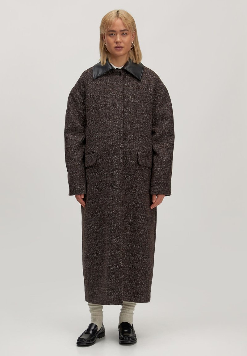 Lang bruine jas van textuurstof, met een zwarte faux leren kraag, twee voorzakken en een rechte silhouette. Draagt over crèmekleurige sokken en zwarte schoenen.