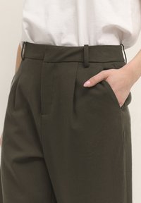Olijfgroene op maat gemaakte broek met een hoge taille, plooien, zijzakken en een gladde stofstructuur. Draagt met een wit shirt.