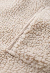 Teksturni bež flis z mehkobno, puhasto površino. Ima majhen bež žep, obrobljen s gladkim robom.