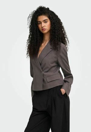 Jeune femme aux longs cheveux bouclés portant un blazer gris ajusté et un pantalon noir ample, mains dans les poches, fond neutre.