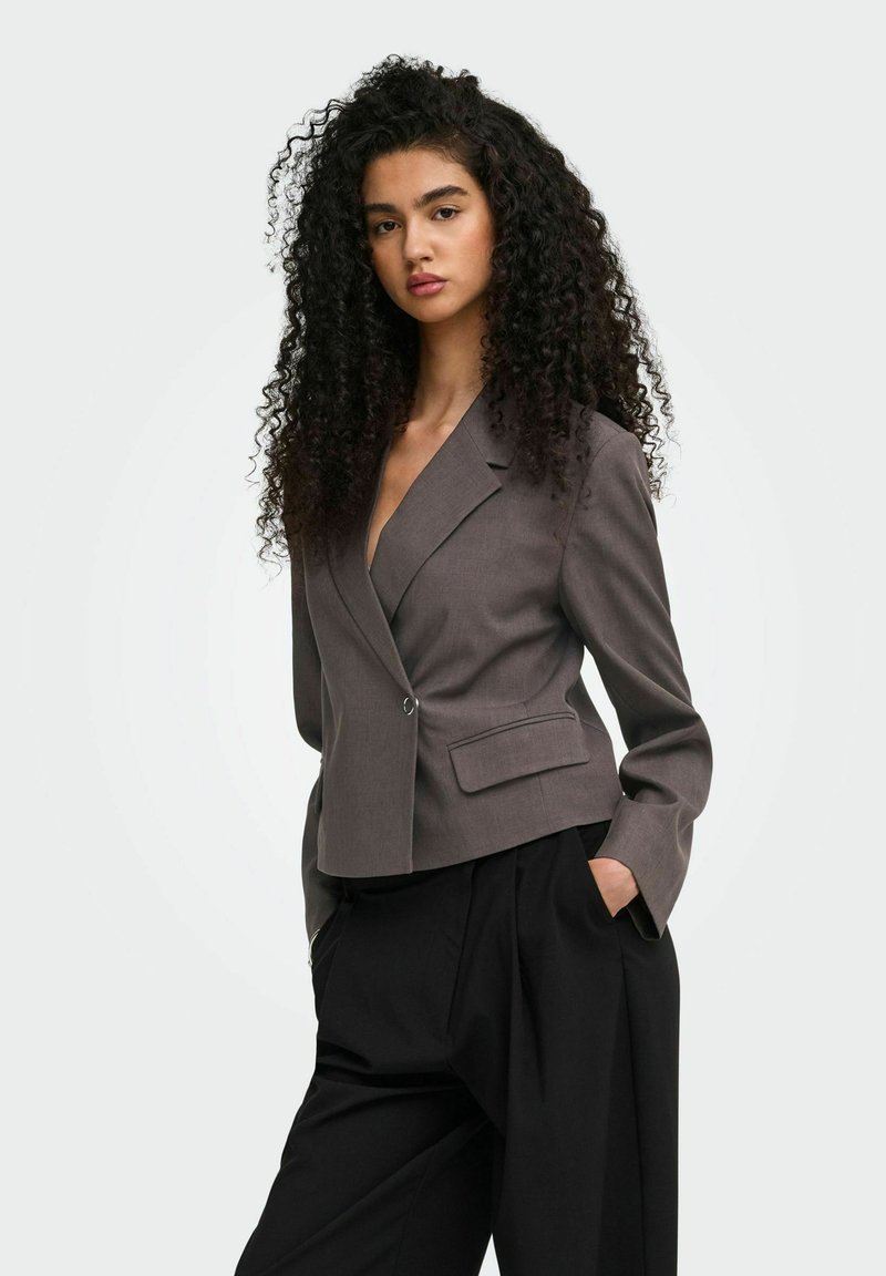 Jeune femme aux longs cheveux bouclés portant un blazer gris ajusté et un pantalon noir ample, mains dans les poches, fond neutre.