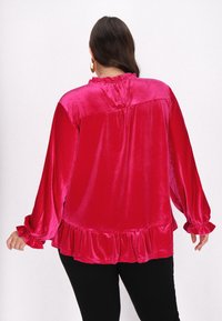 Fuchsia fløjl bluse med lange, puffede ærmer. Har en flæse forneden og en høj halsudskæring med en samlet detalje bagpå.