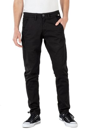 Pantalones de algodón negro con una silueta ajustada, que cuentan con bolsillos laterales, cierre de botón y se combinan con zapatillas negras. Textura y costura visibles.