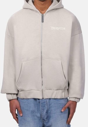 Mann trägt einen hellbeigen Kapuzenpullover mit Reißverschluss und Fronttasche, mit dem Text "Dropstee" auf der linken Brustseite, kombiniert mit blauen Jeans.