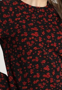 Tissu noir avec un motif dense de petites fleurs rouges ; encolure ronde ; texture légère et transparente ; accents floraux avec des formes de fleurs variées.