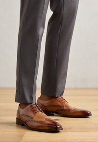 Scarpe eleganti in pelle marrone con design brogue, caratterizzate da perforazioni, finitura lucida e suola nera, abbinate a pantaloni grigi.