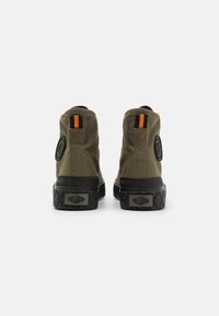 Pantofi sport înalți din pânză verde măsliniu cu tălpi din cauciuc negre, având accente portocalii și o emblema rotundă pe călcâi.