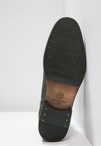 Musta nahkainen kenkäpohja, jossa on teksturoitu pinta ja musiikkinuotteja. Varressa on messinkivärisiä niittejä ja brändilogo. Koko 40.