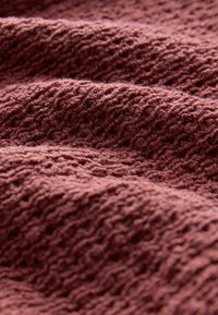 Gestructureerde, geribbelde stof in een diepe bordeauxkleur, met zachte, golvende lagen die de warmte en tactile kwaliteit van het materiaal benadrukken.