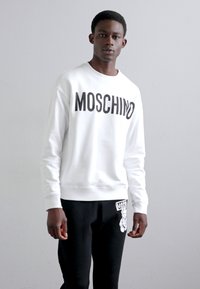 Sudadera blanca con el logo "MOSCHINO" en negro intenso. Combinada con pantalones negros que presentan un diseño gráfico. Material de algodón, estilo minimalista.