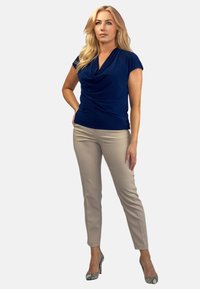 Granatowa bluzka z kołnierzem typu cowl, wykonana z gładkiego materiału, zestawiona z jasnobeżowymi spodniami szytymi na miarę. Modelka ma na sobie wzorzyste szpilki.