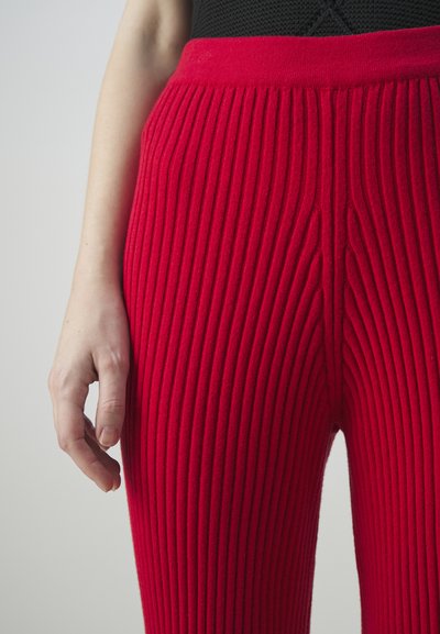 Rote, gerippte Hose aus einem weichen Material, mit einem breiten Bund und vertikalen Strukturmustern. Die Hand ist auf dem Oberschenkel positioniert.