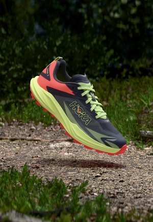 Běžecká trailová bota s neonově žlutou podrážkou, černým a oranžovým svrškem, ve vzduchu nad prašnou cestou s trávou a rozostřeným zeleným pozadím listí.