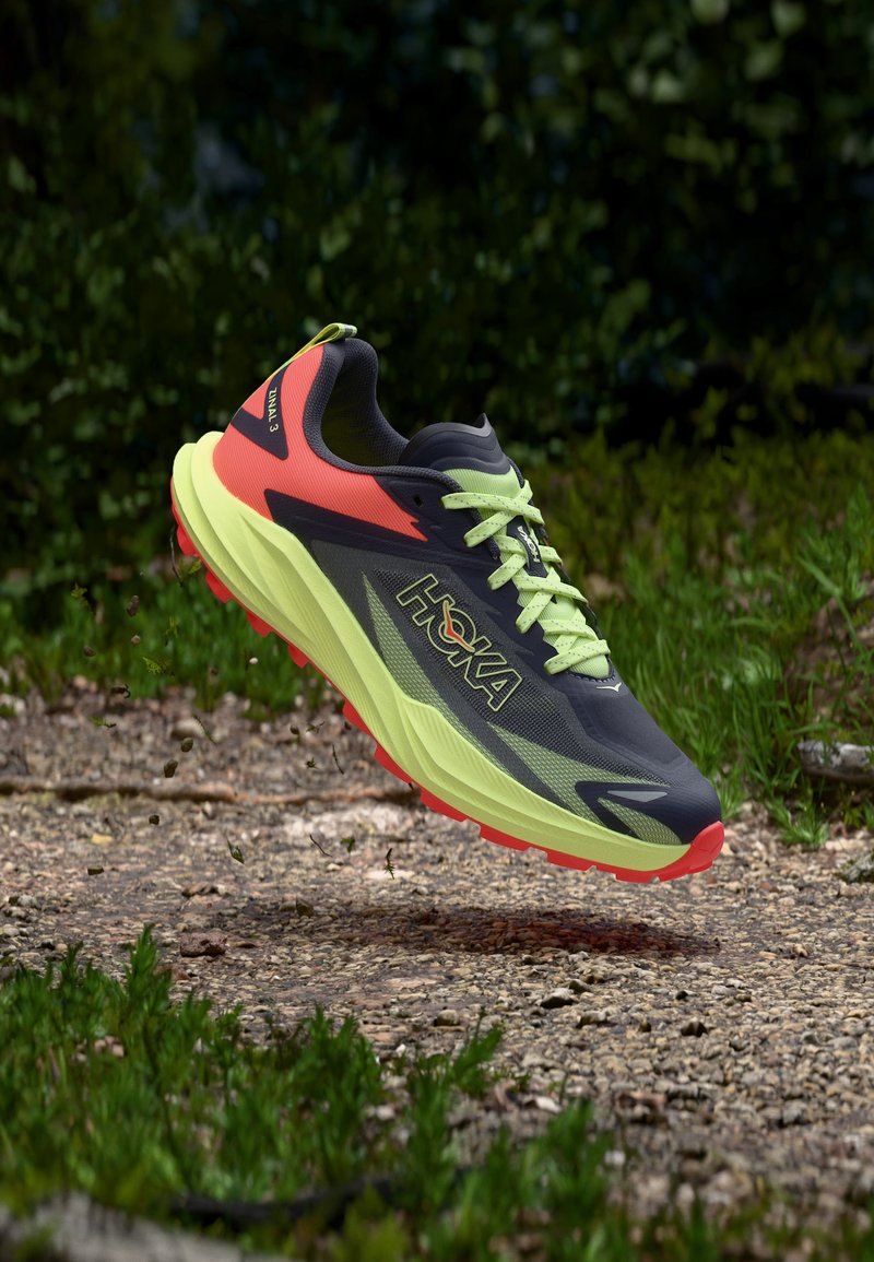 Παπούτσι για τρέξιμο σε μονοπάτι με νέον κίτρινη σόλα, μαύρο και πορτοκαλί επάνω μέρος, στον αέρα πάνω από χωμάτινο μονοπάτι με γρασίδι και θολό πράσινο φόντο φύλλωσης.