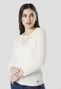 Witte gebreide vrouwen trui met een kanten inzetstuk bij de halslijn, lange mouwen en een aansluitende geribbelde textuur. Gecombineerd met een blauwe spijkerbroek.