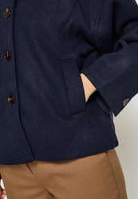 Meresinine villasegu jakk tekstureeritud viimistlusega, millel on suured pruunid nööbid ja küljetasku. Mugav lõige pööratud varrukaservadega.