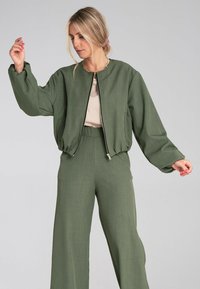 Zielona krótka bomberka z zapięciem na zamek, okrągłym dekoltem i bufiastymi rękawami, zestawiona z pasującymi spodniami o szerokich nogawkach. Gładka tekstura.