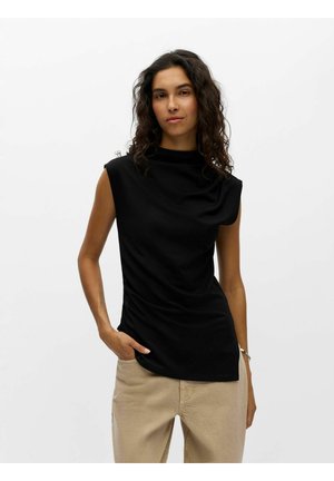 Femme aux cheveux bouclés portant un haut noir sans manches et un pantalon beige, debout avec une main dans la poche devant un fond uni.