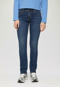 Chude dżinsy w ciemnoniebieskim denimu, z pięcioma kieszeniami, ozdobnymi szwami, noszone z srebrnymi butami sportowymi.