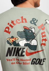 Kreslený aligátor v golfovém klobouku, který drží pizzu a golfovou hůl, obklopený textem: "Pitch & Putt," "NIKE," "Na našem výkonu budete naladěni!" a "GOLF."
