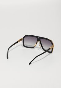 Occhiali da sole neri oversize con lenti sfumate, dettagli in tonalità oro e design angolare. Tempi neri eleganti e montatura prominente sono notevoli.