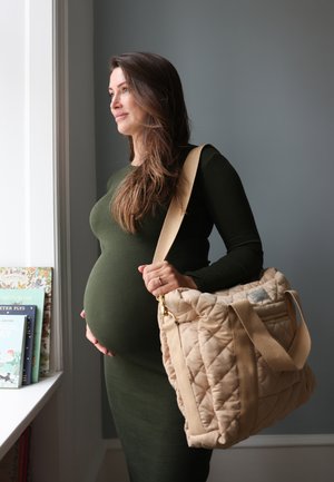 Tehotná žena v tmavozelených šatách stojí pri okne, drží na ramene svetlohnedú prešívanú tašku a rukou si hladí tehotenské bruško.