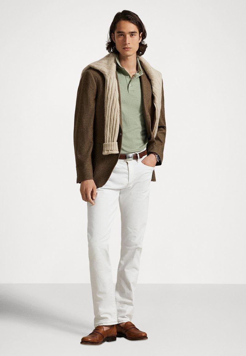 Brauner Tweed-Blazer, hellgrünes Polohemd, cremefarbener Zopfpullover über den Schultern gelegt, weiße Hose und braune Lederschuhe.