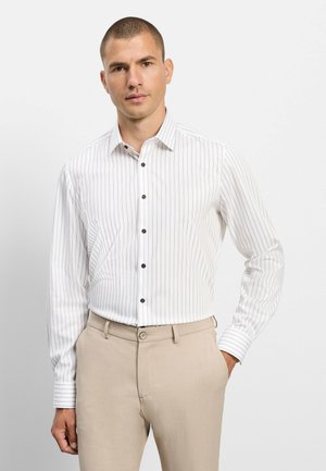 Mann trägt ein weißes Langarmhemd mit dünnen vertikalen Streifen, beige Hose, steht mit einer Hand in der Tasche vor weißem Hintergrund.