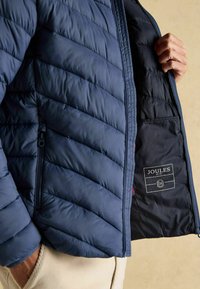 Marineblå polstret jakke med et quiltet design, der har en sidelomme med lynlås og et indre etiket med mærket "Joules."