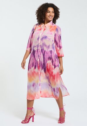 Kobieta o kręconych włosach, ubrana w kolorową sukienkę midi na guziki, z długim rękawem, z wzorem tie-dye i różowe sandały na wysokim obcasie.