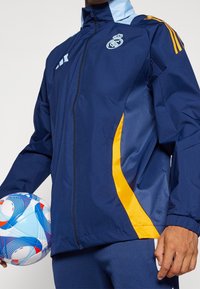 Donkerblauwe jas met lichtblauwe accenten, voorzien van een ritssluiting aan de voorkant, zijzakken en oranje strepen. Houdt een meerkleurige voetbal vast.