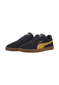 Une paire de baskets basses en daim bleu marine de Puma avec des rayures jaune moutarde et des semelles en caoutchouc marron, lacées, légèrement inclinées vers la gauche.