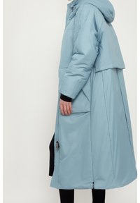 Cappotto lungo imbottito azzurro chiaro con cappuccio, dotato di chiusura con zip, tasche laterali e vestibilità morbida; il tessuto appare liscio e leggermente riflettente.