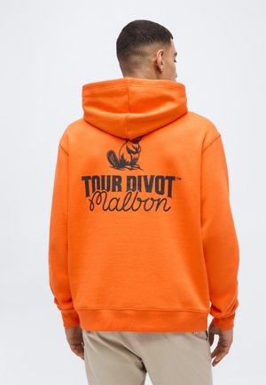 Homme de dos portant un sweat à capuche orange vif avec un logo de castor noir et le texte "TOUR DIVOT Malbon" au dos, pantalon beige.
