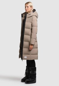 Langer beiger Puffer-Mantel mit Kapuze, ausgestattet mit einem schwarzen Reißverschluss und gepolsterten Abschnitten. Schwarze wasserfeste Stiefel mit Schnürsenkeln und strukturierten Details.