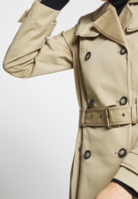 Abrigo trench beige hecho de tela suavemente, con cintura ceñida con cinturón, botones de doble botonadura y detalles de costura en contraste.