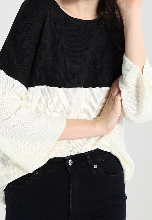 Donna che indossa un maglione lavorato a maglia a maniche lunghe bianco e nero con maniche a campana e jeans neri, braccia incrociate in vita.