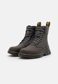 Paire de bottes montantes gris foncé avec semelles épaisses noires, lacets blancs et noirs, coutures jaunes, et languettes de traction jaunes à logo à l'arrière.