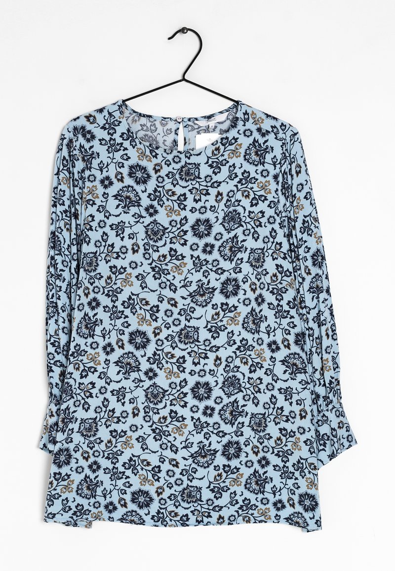 Blouse bleu clair à manches longues avec col rond, présentant un motif floral bleu marine et beige, suspendue sur un cintre noir sur fond blanc.