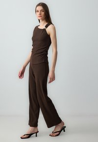 Débardeur marron texturé avec un détail froncé et un accent doré sur l'épaule ; associé à un pantalon large marron et des sandales à talons noires.