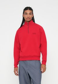 Sudadera roja con cremallera y cuello alto, puños y bajo acanalados, con un pequeño logo negro en el pecho y textura suave.