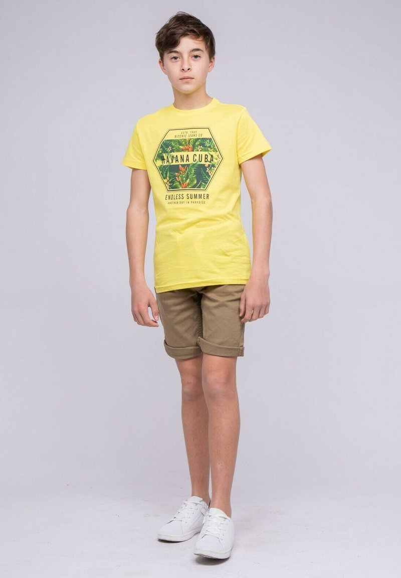 T-shirt en coton jaune avec un graphique tropical hexagonal, associé à un short beige. Baskets blanches à semelle plate. Design simple et décontracté.
