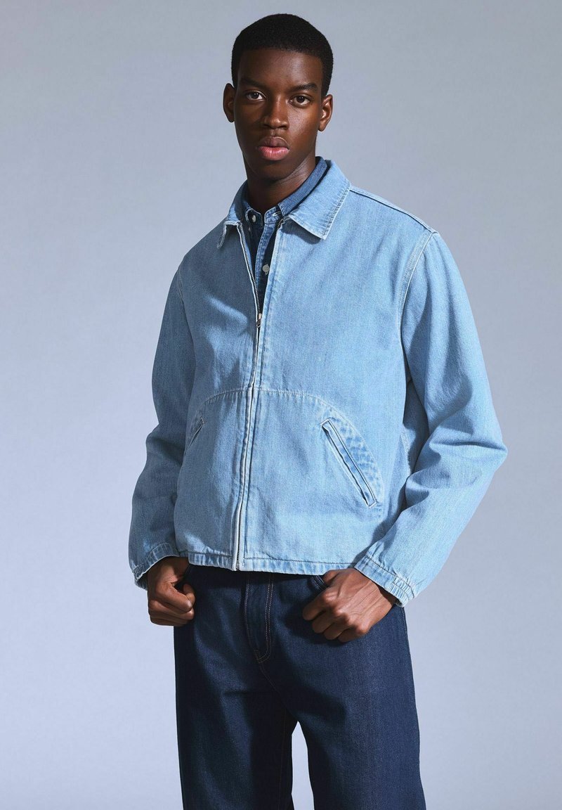 Helle blaue Jeansjacke mit Reißverschluss vorne, taillierter Passform und zwei seitlichen Taschen. Lange Ärmel mit klassischem Kragen und minimalen Details.