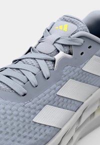 Baskets de sport grises en tissu texturé, accents blancs et jaunes, design à lacets et rayures blanches proéminentes sur le côté.
